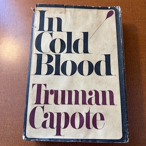May include: A hardcover book titled In Cold Blood by Truman Capote. The cover features a cream-colored background with bold black lettering for the title and dark purple lettering for the author's name, accented by a small purple spoon graphic.