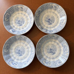 Può includere: Un set di quattro ciotole rotonde in ceramica con un motivo a trasferimento blu e bianco. Ogni ciotola mostra una scena pastorale centrale con una persona seduta in un paesaggio, circondata da un bordo floreale e vignette rettangolari.