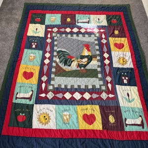 Puede incluir: Una colcha de patchwork rectangular que presenta un panel central con una ilustración de un gallo. El borde consta de cuadrados que muestran varios motivos, incluyendo manzanas, tazas de café, soles, corazones, camas y tulipanes con texto.