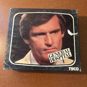 May include: A square cardboard game box featuring a portrait of a man with brown hair wearing a light-colored shirt. The box is black with a white border and includes the text GENERAL HOSPITAL in white letters and the brand name TUCO in the corner.