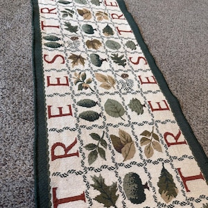 May include: A rectangular table runner featuring a cream-colored background with a grid pattern. The design includes various green and brown leaf and tree illustrations within the grid squares, with the word TREES printed in red along the sides.