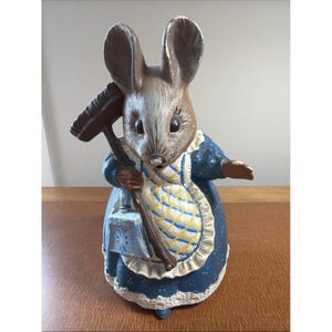 May include: A painted ceramic figurine of a mouse standing upright, wearing a dark blue dress with white polka dots and a yellow and white checkered apron. The mouse holds a brown broom and a small blue dustpan, featuring a white lace trim.
