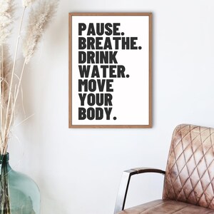 May include: A rectangular wall art print in a light brown wooden frame featuring black sans-serif text on a white background that reads: PAUSE. BREATHE. DRINK WATER. MOVE YOUR BODY. The print is displayed on a plain white wall above a brown chair.