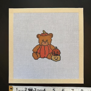 Op de afbeelding: Een vierkant borduurcanvas met een bruine teddybeer in een pompoenkostuum met bijpassende hoed, zittend naast twee kleine pompoenlantaarns. Een Dritz-meetlint toont aan dat het canvas een breedte heeft van ongeveer 20,3 cm.