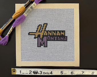May include: A cross-stitch project featuring the name "Hannah Montana" in gold and purple thread on a white grid canvas. Two spools of embroidery floss in gold and purple are placed above the canvas. A measuring tape shows the canvas is approximately 5 inches wide.