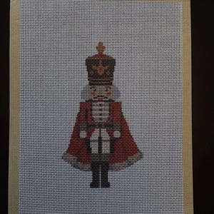 May include: A needlepoint canvas featuring a nutcracker soldier design. The figure wears a tall red and brown hat, a red coat with white detailing, and black boots. The design is centred on a white grid canvas with a tan border tape on all sides.