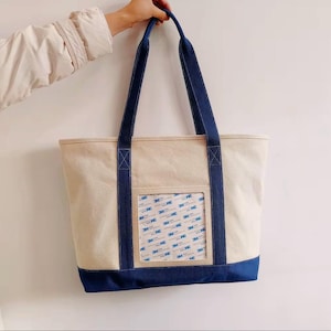 Op de afbeelding: Een beige canvas tote bag met marineblauwe accenten. De tas heeft marineblauwe handvatten en een marineblauwe bodem. Een doorzichtige zak aan de voorkant toont een patroon van 3M-stickers. De tas wordt door een persoon vastgehouden.
