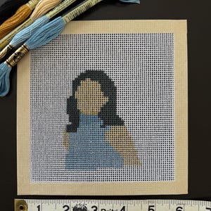 May include: A cross-stitch portrait of a person with dark hair and a blue top. The artwork is on a white grid canvas with a beige border. Several spools of embroidery floss in various colours are arranged above the canvas. A measuring tape is at the bottom.
