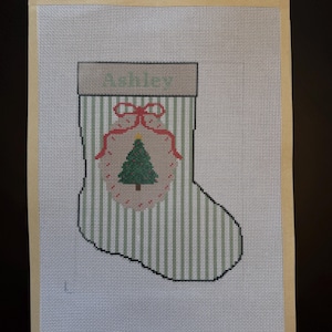 May include: A needlepoint canvas featuring a Christmas stocking design with green and white vertical stripes, a tan cuff, and a central ornament graphic containing a green pine tree. A measuring tape shows the canvas width is 13 inches.