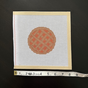 May include: A square needlepoint canvas featuring a circular waffle design in shades of brown and orange. The canvas is bordered by tan tape and measures approximately 23 cm in width as shown by the Dritz measuring tape placed below it.