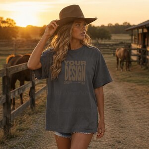 Może przedstawiać: Ciemnoszary t-shirt z krótkim rękawem z napisem YOUR DESIGN nadrukowanym w beżowym kolorze na klatce piersiowej. Koszulka ma luźny, swobodny krój z okrągłym dekoltem i jest noszona z brązowym kapeluszem z szerokim rondem.