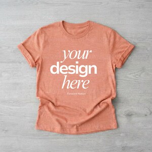 May include: A peach-coloured t-shirt with rolled-up sleeves. The shirt features the text "your design here" in white, with "Forward Notion" below. The shirt is laid flat on a grey wooden surface.