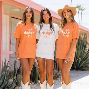May include: Three women wearing oversized t-shirts with the text "your design here" in white. Two shirts are orange, and one is white. They are wearing white cowboy boots. The background includes a pink building and cacti.