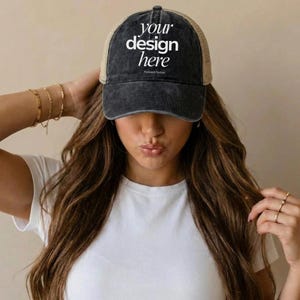 Könnte beinhalten: Eine schwarze und beige Trucker-Cap mit dem weißen Text "your design here". Die Kappe hat einen Used-Look und eine Mesh-Rückseite. Die Kappe wird von einer Person mit langen braunen Haaren getragen.