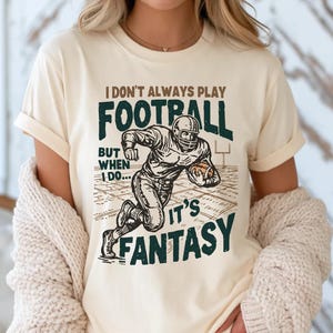 May include: A cream-colored short-sleeve t-shirt featuring a graphic of a football player running with a ball. The text reads: I DON'T ALWAYS PLAY FOOTBALL BUT WHEN I DO... IT'S FANTASY. The design uses dark green and brown ink colors.