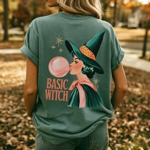 May include: A sage green t-shirt featuring a graphic on the back of a witch wearing a pointed hat with a leopard print band, blowing a pink bubblegum bubble. The text "BASIC WITCH" is printed in coral pink below the illustration with star accents.