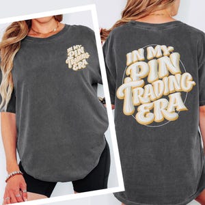 May include: A charcoal gray short-sleeve t-shirt featuring the text "IN MY PIN TRADING ERA" in a large, cream-colored, bubble-style font with a thin yellow outline on the back and a smaller version of the same text on the front left chest.