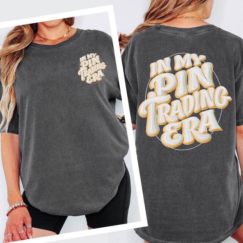 May include: A charcoal gray short-sleeve t-shirt featuring the text "IN MY PIN TRADING ERA" in a large, cream-colored, bubble-style font with a thin yellow outline on the back and a smaller version of the same text on the front left chest.