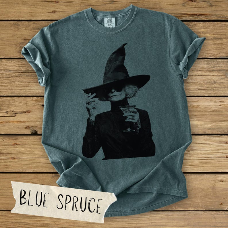 Puede incluir: Una camiseta de manga corta color azul abeto con un estampado gr&aacute;fico negro de una persona con un sombrero de bruja de ala ancha y gafas de sol, sosteniendo una copa de martini. La prenda tiene cuello redondo y una etiqueta de marca visible.