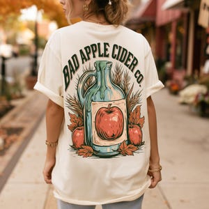 Puede incluir: Una camiseta color crema con un gráfico en la espalda de una jarra de cristal con líquido, una manzana roja grande y tallos de trigo. El texto "BAD APPLE CIDER CO." se arquea sobre la ilustración, decorada con hojas de otoño.