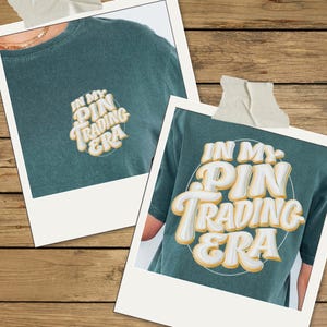May include: A dark teal t-shirt featuring the text "IN MY PIN TRADING ERA" in a bold, white, and yellow bubble-style font. The design is displayed in two sizes, with a smaller version on the chest and a larger version on the back of the shirt.