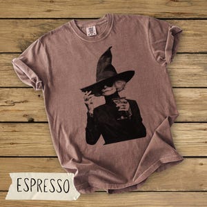 Puede incluir: Una camiseta de cuello redondo color rosa empolvado con un estampado gr&aacute;fico en blanco y negro de una persona con un sombrero de bruja de ala ancha y gafas de sol, sosteniendo un cigarrillo y una copa de c&oacute;ctel en las manos.
