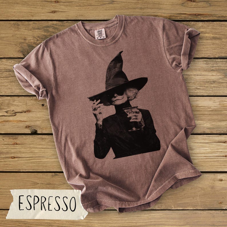 Puede incluir: Una camiseta de cuello redondo color rosa empolvado con un estampado gr&aacute;fico en blanco y negro de una persona con un sombrero de bruja de ala ancha y gafas de sol, sosteniendo un cigarrillo y una copa de c&oacute;ctel en las manos.