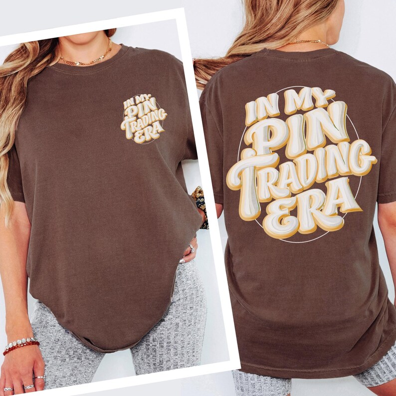 May include: A dark brown short-sleeve t-shirt featuring the text "IN MY PIN TRADING ERA" printed in a white and yellow layered font on the left chest and in a larger version on the back, enclosed within a thin white circular outline.