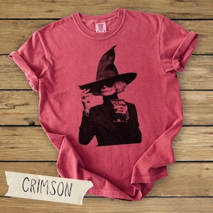 Puede incluir: Una camiseta roja de manga corta con un estampado gr&aacute;fico negro de una persona con un sombrero de bruja de ala ancha y gafas de sol, sosteniendo una copa de c&oacute;ctel. La prenda tiene cuello redondo y una etiqueta Comfort Colors en el cuello.