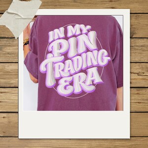 May include: A plum-colored short-sleeve t-shirt featuring a circular graphic on the back with the white and purple text "IN MY PIN TRADING ERA" in a bold, bubbly, retro-inspired font style. The shirt has a relaxed fit and a solid fabric texture.