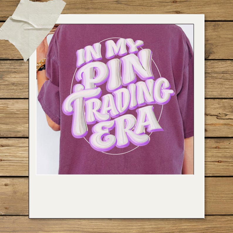 May include: A plum-colored short-sleeve t-shirt featuring a circular graphic on the back with the white and purple text "IN MY PIN TRADING ERA" in a bold, bubbly, retro-inspired font style. The shirt has a relaxed fit and a solid fabric texture.