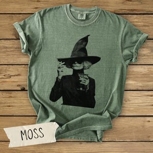 Puede incluir: Una camiseta de cuello redondo color verde salvia con un estampado gr&aacute;fico en blanco y negro de una persona con sombrero de bruja y gafas de sol sosteniendo una copa de c&oacute;ctel. Una etiqueta blanca con la palabra MOSS est&aacute; abajo a la izquierda.
