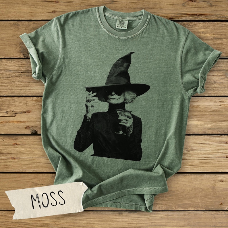 Puede incluir: Una camiseta de cuello redondo color verde salvia con un estampado gr&aacute;fico en blanco y negro de una persona con sombrero de bruja y gafas de sol sosteniendo una copa de c&oacute;ctel. Una etiqueta blanca con la palabra MOSS est&aacute; abajo a la izquierda.