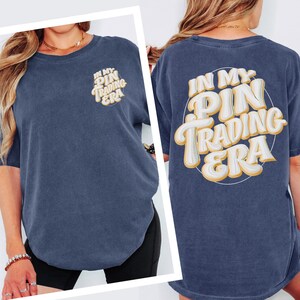 May include: A navy blue short-sleeve t-shirt with a vintage wash. The front left chest features the text "IN MY PIN TRADING ERA" in white and yellow. The back displays the same text in a large, bold, rounded font with a white and yellow gradient.