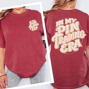 May include: A maroon short-sleeve t-shirt featuring a small graphic on the front left chest and a large graphic on the back. Both graphics display the text "IN MY PIN TRADING ERA" in a stylized, layered white and yellow font within a circle.