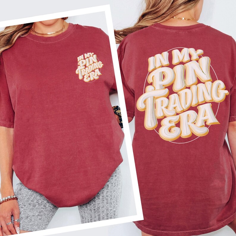 May include: A maroon short-sleeve t-shirt featuring a small graphic on the front left chest and a large graphic on the back. Both graphics display the text "IN MY PIN TRADING ERA" in a stylized, layered white and yellow font within a circle.