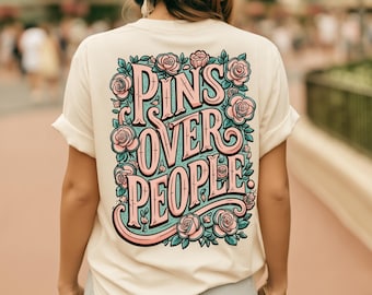 May include: A cream-colored t-shirt featuring a large graphic on the back with the text PINS OVER PEOPLE in stylized pink lettering. The design is surrounded by illustrations of pink roses and teal leaves, creating a decorative floral border.