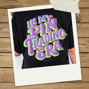 May include: A black t-shirt featuring a graphic design with the text IN MY PIN TRADING ERA in bold, layered purple and lime green lettering. The text is arranged in a circular formation, creating a vibrant, multi-dimensional appearance.