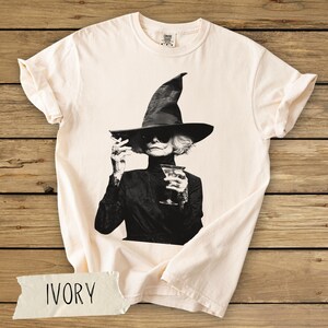 Puede incluir: Una camiseta de manga corta color marfil con un estampado gr&aacute;fico en blanco y negro de una persona con un sombrero de bruja puntiagudo y gafas de sol, sosteniendo un cigarrillo y una copa de martini.