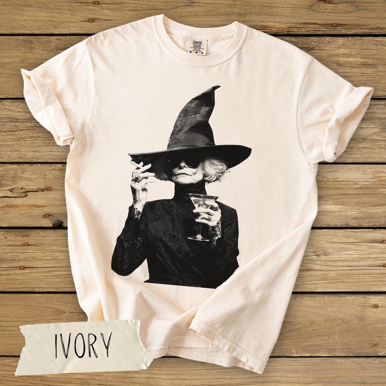 Puede incluir: Una camiseta de manga corta color marfil con un estampado gr&aacute;fico en blanco y negro de una persona con un sombrero de bruja puntiagudo y gafas de sol, sosteniendo un cigarrillo y una copa de martini.