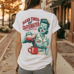 Puede incluir: Una camiseta blanca con el gráfico de una camarera con aspecto de zombi en uniforme azul verdoso, vertiendo café de una jarra de cristal en una taza roja. El texto sobre la ilustración dice "DEAD TIRED BUT CAFFEINATED" en letras rojas y azul verdosas.