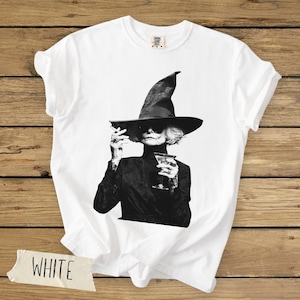 Puede incluir: Una camiseta blanca de manga corta con un estampado gr&aacute;fico en blanco y negro de una persona con un sombrero de ala ancha y gafas de sol, sosteniendo un cigarrillo y una copa de martini. La camiseta tiene una etiqueta de Comfort Colors.