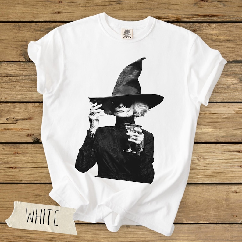 Puede incluir: Una camiseta blanca de manga corta con un estampado gr&aacute;fico en blanco y negro de una persona con un sombrero de ala ancha y gafas de sol, sosteniendo un cigarrillo y una copa de martini. La camiseta tiene una etiqueta de Comfort Colors.