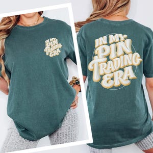 May include: A dark teal short-sleeve t-shirt featuring the text IN MY PIN TRADING ERA in a large, cream-colored, bubble-style font on the back and a smaller version of the same text on the front left chest area.