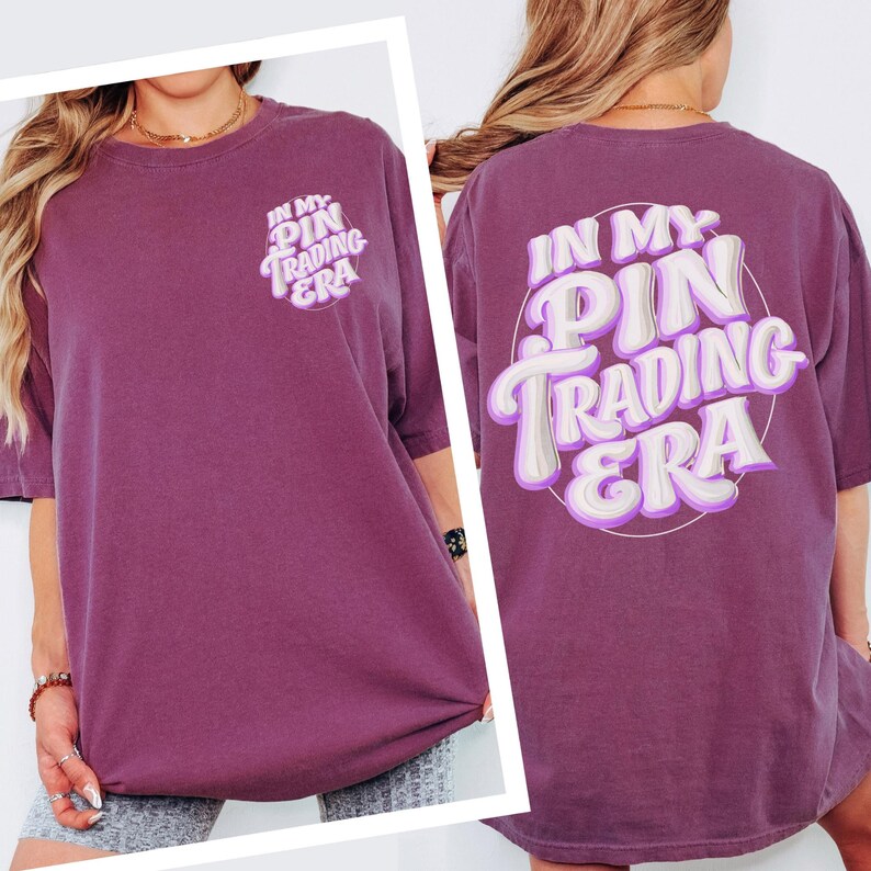 May include: A plum-colored short-sleeve t-shirt featuring the text "IN MY PIN TRADING ERA" printed in a white and light purple layered font. The front displays a small version of the text, while the back features a large version of the same design.