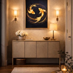May include: A framed artwork featuring two golden koi fish swimming in a circular pattern against a dark background. The artwork hangs above a light-colored cabinet with a vase of white flowers, a lamp, and a decorative bowl.