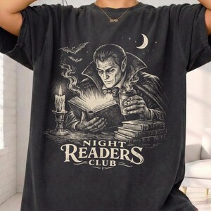 以下が含まれることがあります： 黒と白のイラストが描かれたダークグレーのTシャツ。吸血鬼が本を読んでおり、ろうそく、コウモリ、三日月が描かれています。「NIGHT READERS CLUB」の文字がイラストの下にあります。