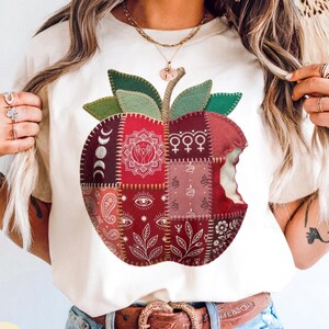 Op de afbeelding: Ivoorkleurig T-shirt met een patchwork appelontwerp. De appel bestaat uit rode, bordeauxrode en roze stoffen vierkanten met hemelse en bloemmotieven. De appel heeft een hap uit en groene bladeren en een steel maken het ontwerp compleet.