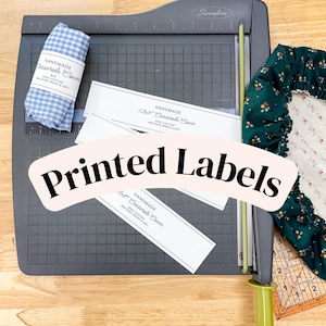 May include: A dark gray Swingline paper cutter with a green handle, featuring white paper labels with black text reading Handmade Casserole Cover, 100% Cotton, Machine Wash & Dry, alongside a blue gingham fabric roll and a dark green floral fabric.