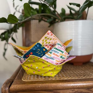 May include: A stack of colorful fabric bowls with various fruit and vegetable patterns. The bowls are yellow, pink, and teal, with designs of strawberries, bananas, and other produce. The bowls are stacked on a wooden table.